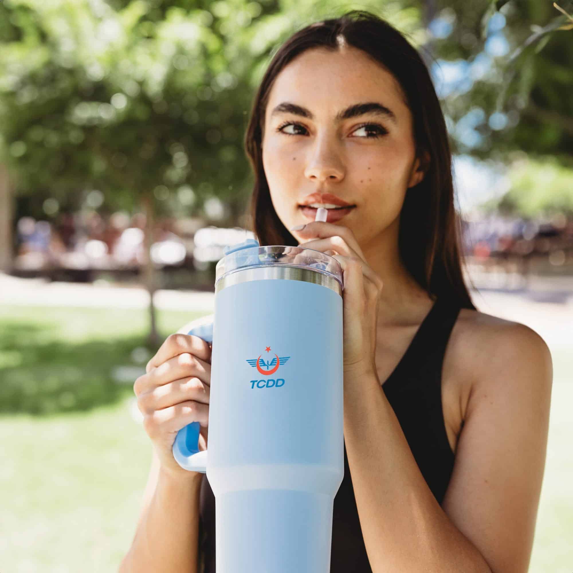 A drink bottle in light blue with a handle, featuring a logo and a clear lid, held by a person outdoors.