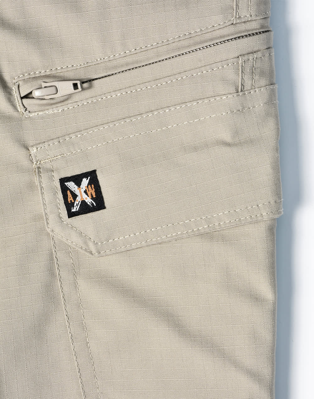 A close-up of light khaki work pants featuring a zippered pocket and a small logo on the side.