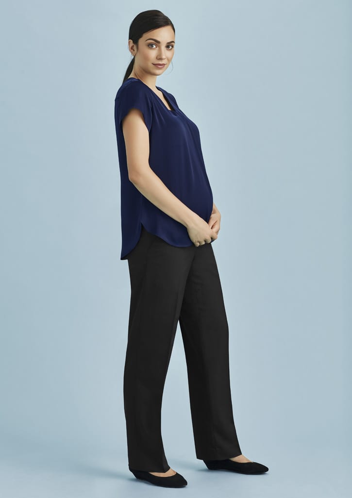 A woman wearing a navy top and black maternity pants stands against a light blue background, smiling and holding her belly.