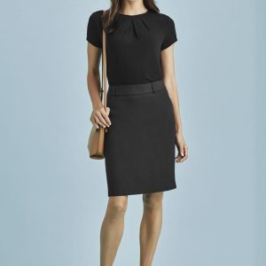 A woman stands wearing a black multi-pleat skirt with a matching top, complemented by beige heels and a handbag.