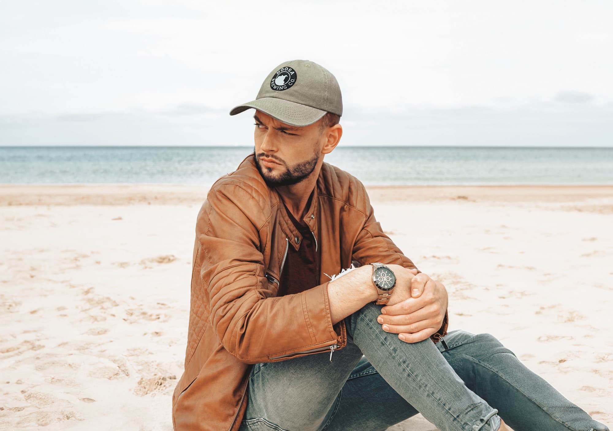A man with a cap sits on the beach, wearing a brown jacket and grey jeans. The cap is olive green with a logo.