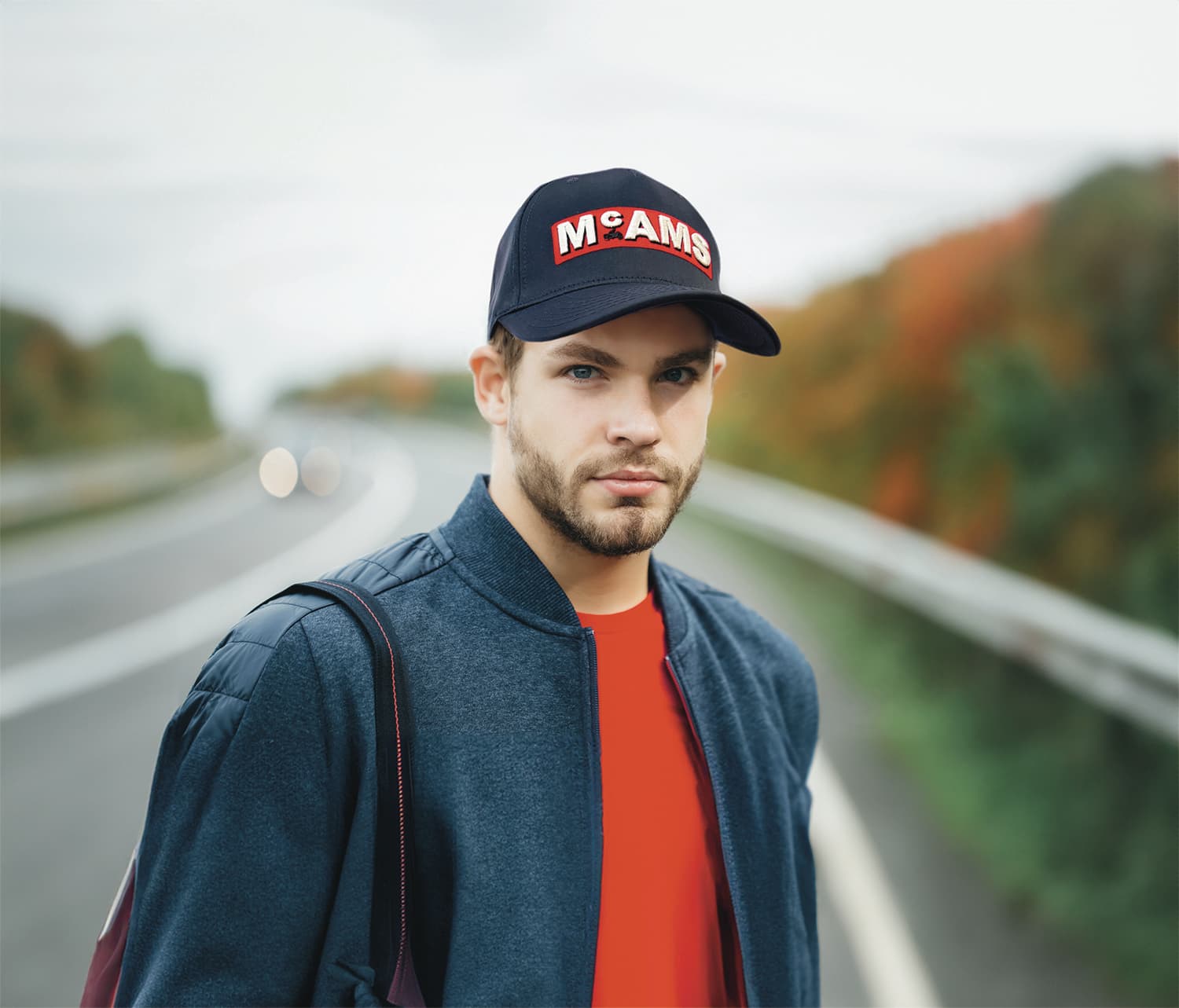 A navy twill cap with a logo, worn by a man in a grey jacket, standing on a road with autumn foliage in the background.