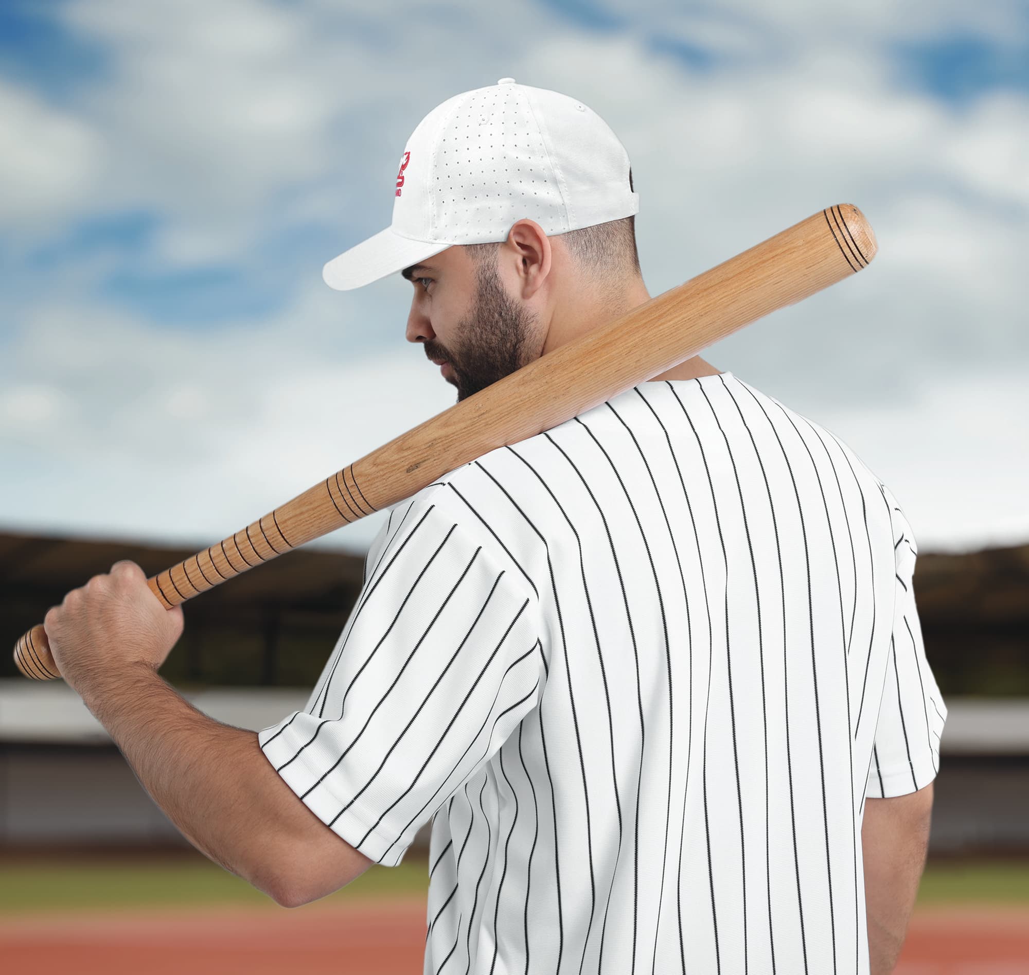 A man in a striped baseball jersey holds a wooden bat while wearing a white breathable cap with perforations.