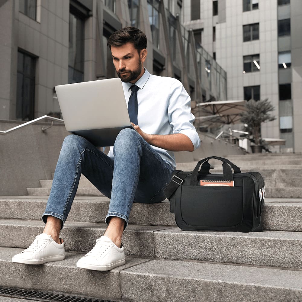 A black laptop briefcase sits on steps beside a man working on a silver laptop, dressed in a white shirt and tie.
