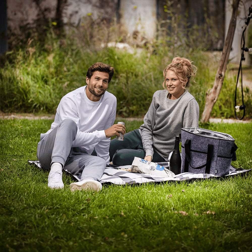 A drink bottle cooler bag in grey felt, surrounded by two people enjoying snacks on a picnic blanket in a grassy area.