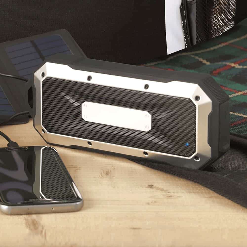 A waterproof outdoor Bluetooth speaker in black and silver, placed next to a smartphone on a wooden surface.