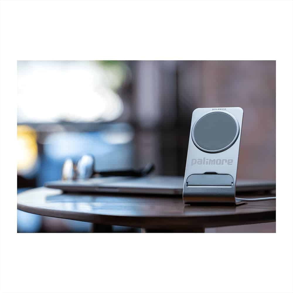A silver wireless charging stand rests on a wooden table beside glasses and a laptop, featuring a logo.