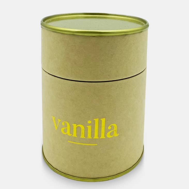A small, round candle in a brown container with a gold lid, featuring 