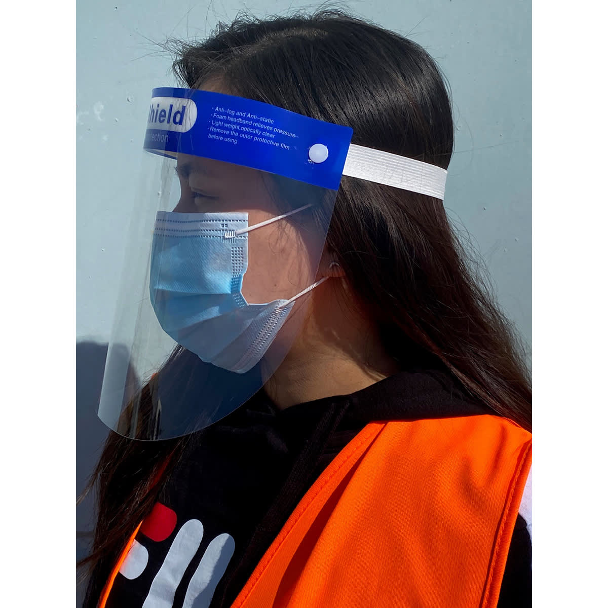 A clear full-length face shield worn by a person with a blue headband and an orange safety vest, featuring a logo.