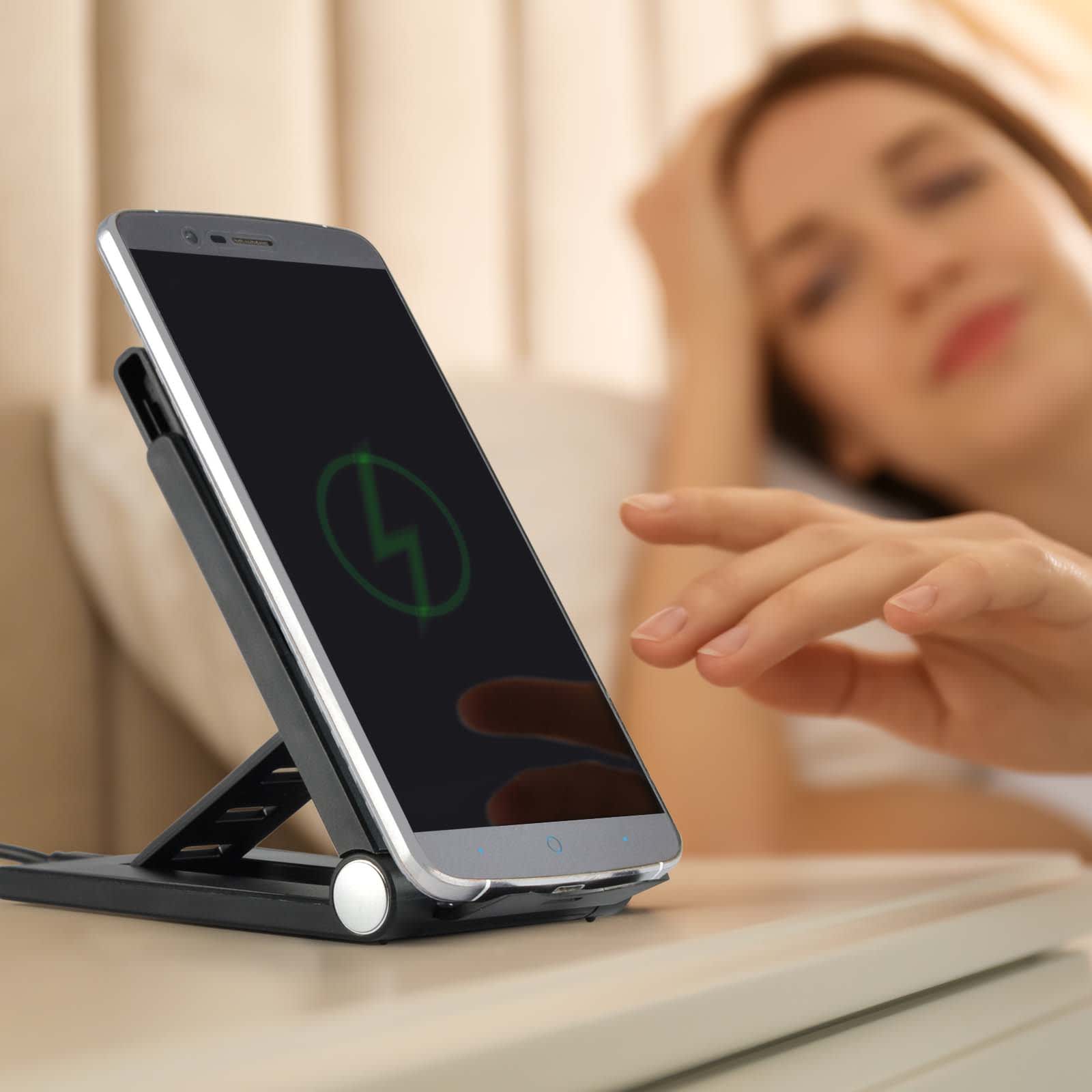 A black wireless charger on a bedside table with a smartphone resting on it, featuring a person in the background.