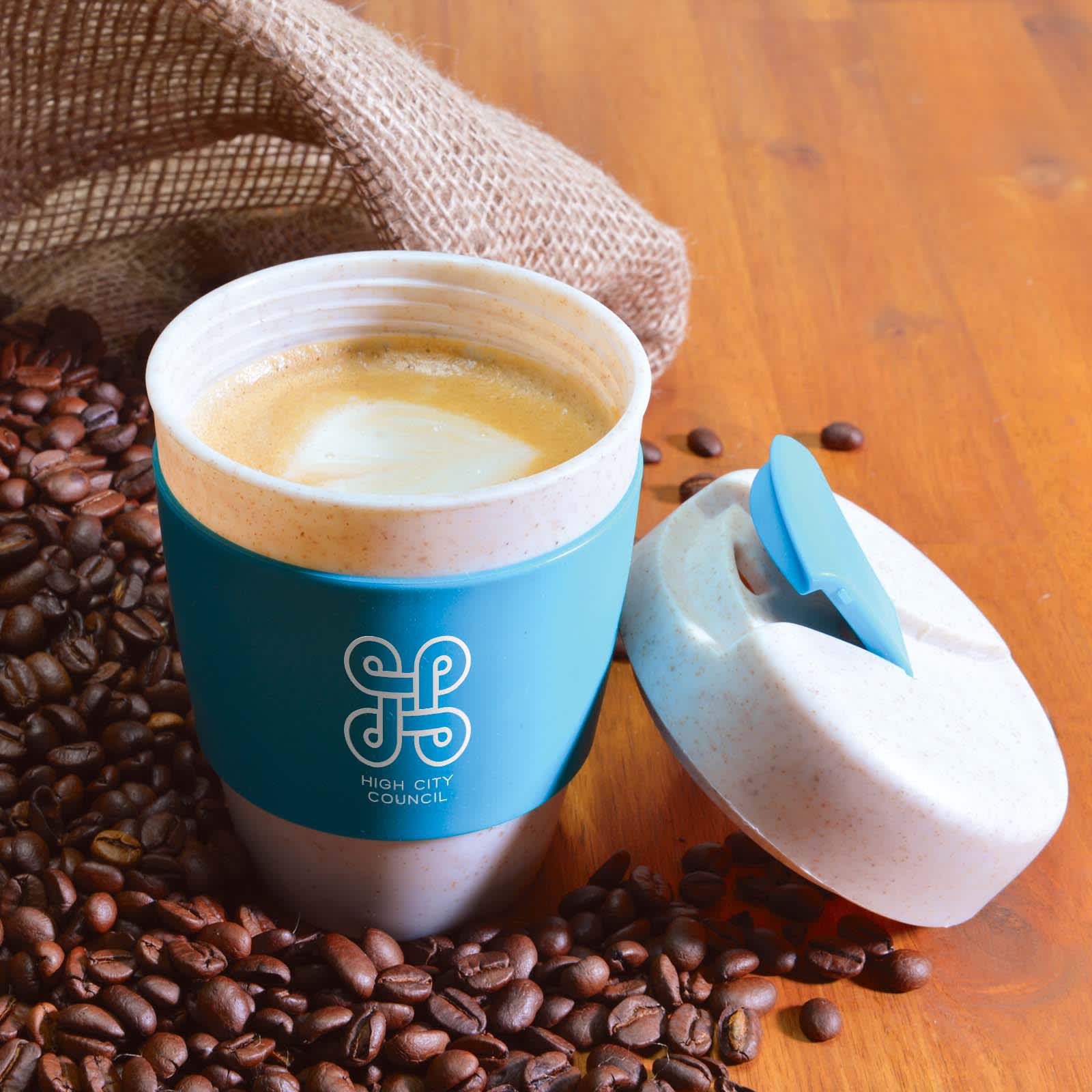 A reusable coffee cup with a blue silicone band, surrounded by coffee beans on a wooden surface.