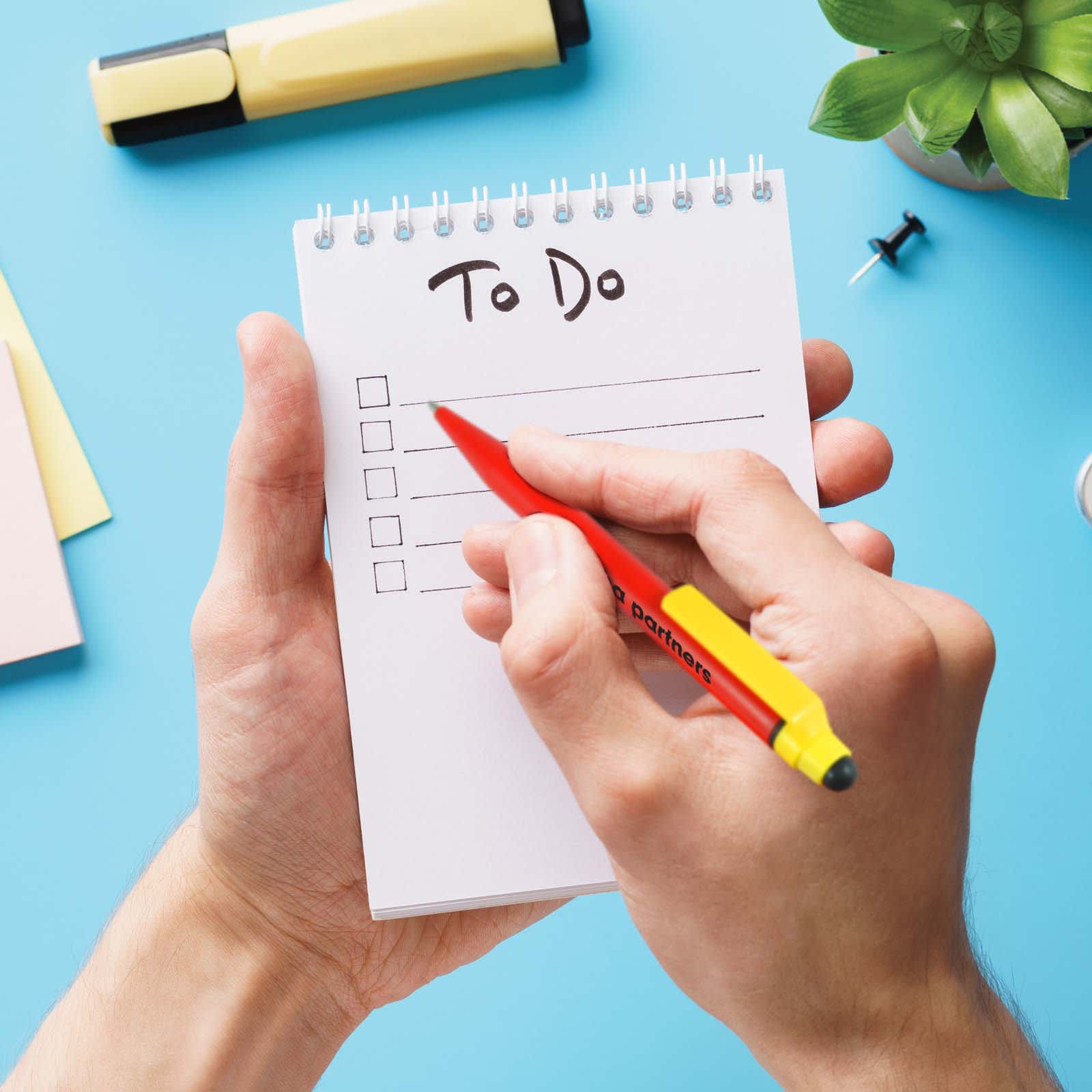 A hand holding a red Chameleon pen writing on a notepad with a 