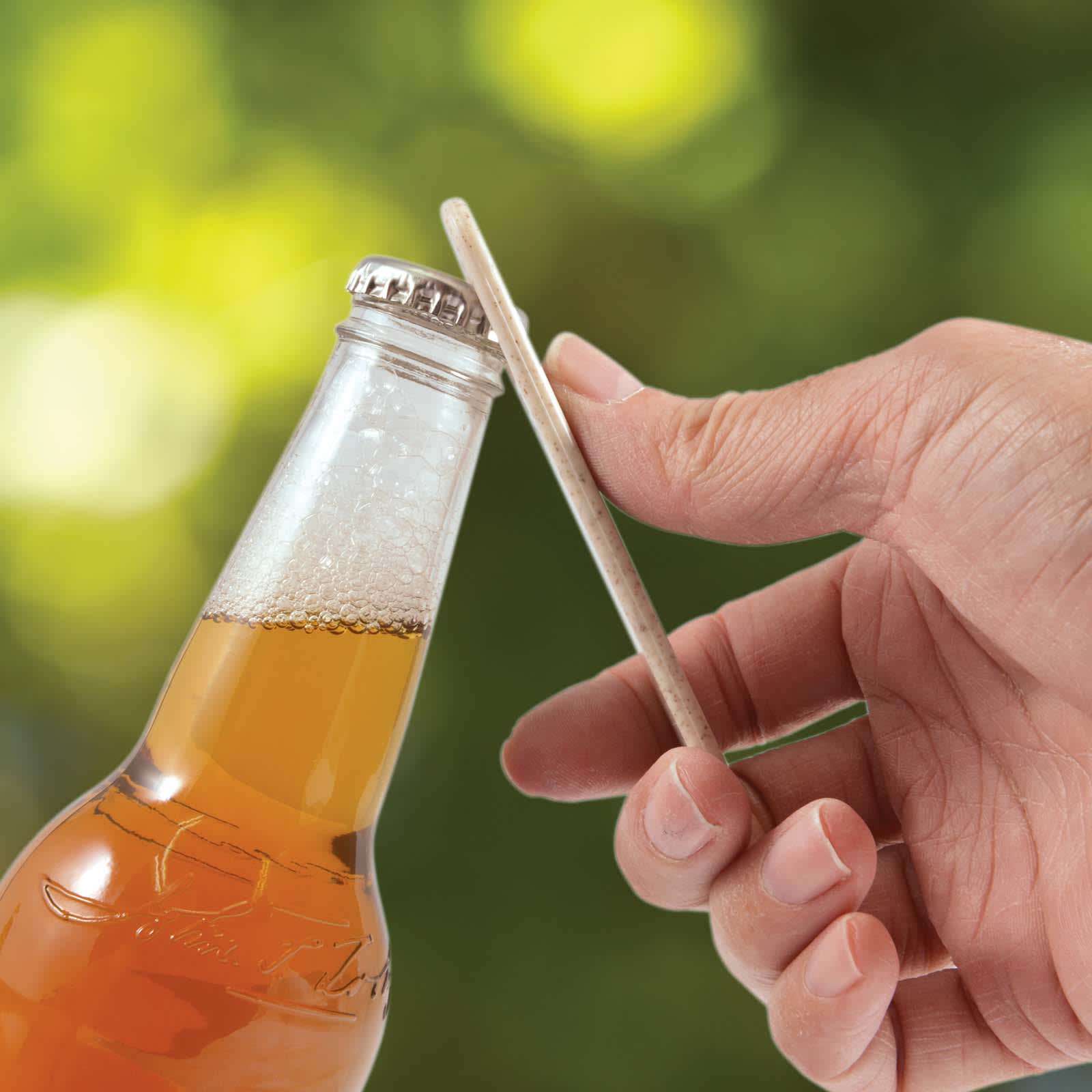 A drink bottle with a gold cap is being opened using a white Arctic Bottle Opener, set against a blurred green background.