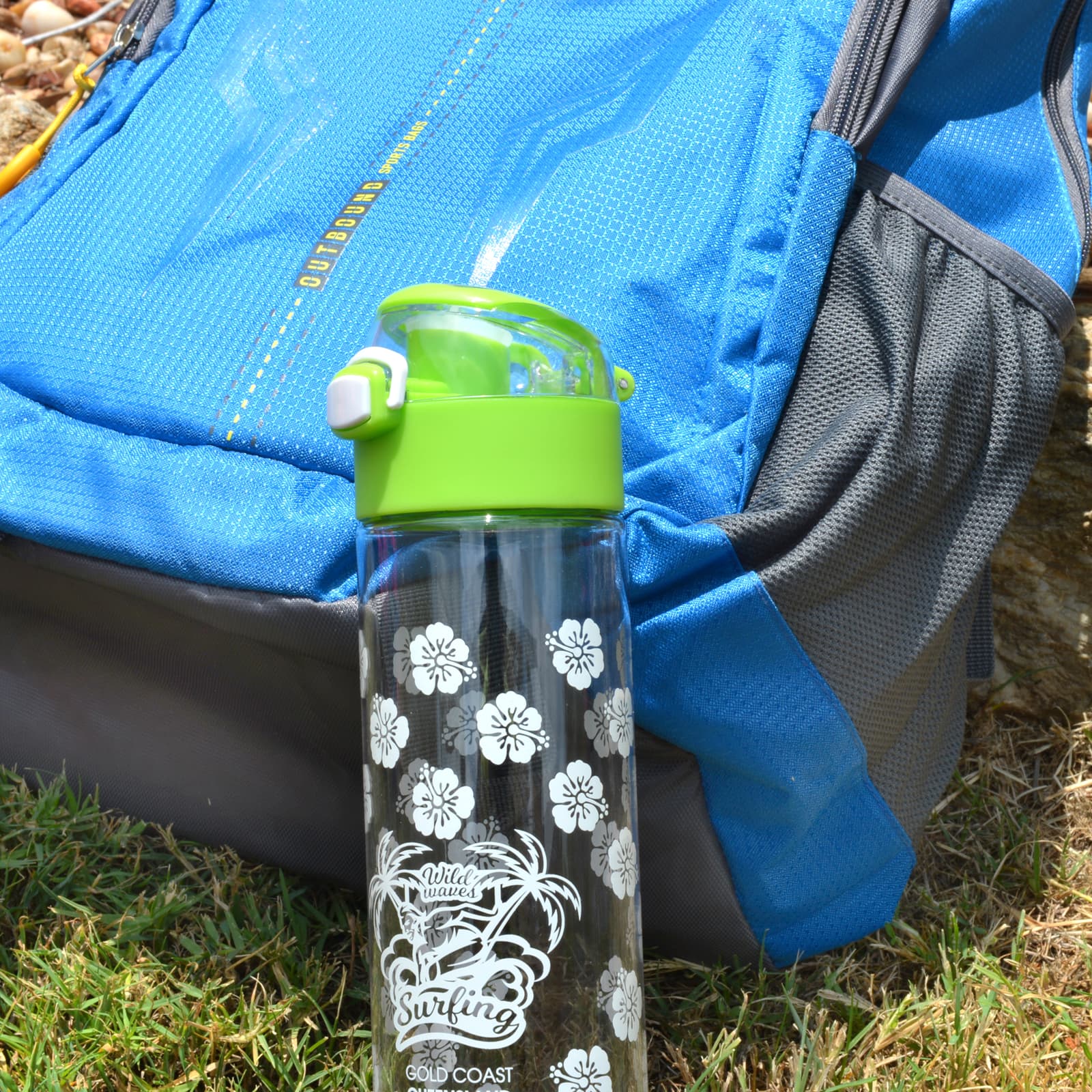 A drink bottle with a clear body featuring white floral designs and a green flip cap, placed next to a blue backpack.