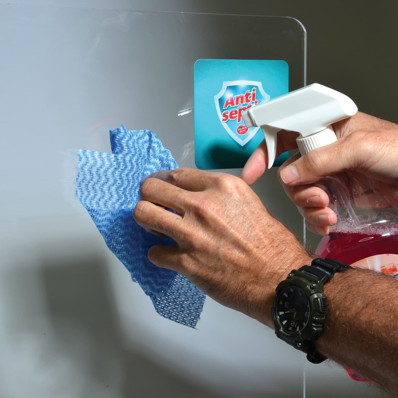 A clear personal protection screen with a hand holding a spray bottle and a blue cleaning cloth.