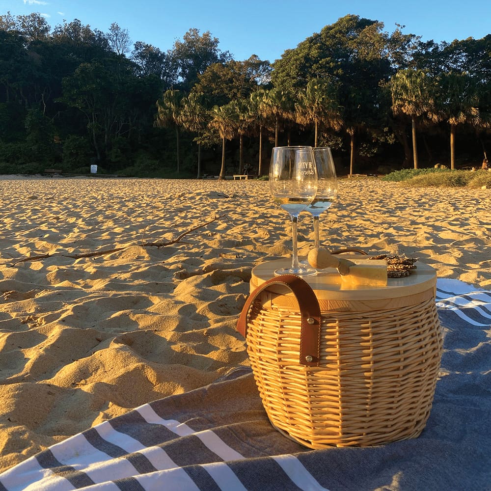 Wicker cooler basket in natural colour with a wooden lid, featuring crackers, grapes, and a drink bottle.