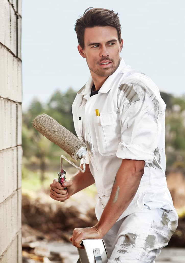 A man in a white cotton drill overall stands with a paint roller, set against a blurred outdoor background.