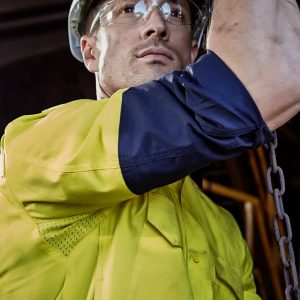 A rugged long sleeve shirt in yellow and navy, featuring reflective elements and a logo, worn by a person in safety gear.