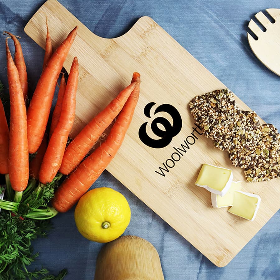 A bamboo chopping board featuring a logo, with fresh carrots, a lemon, and slices of cheese on display.