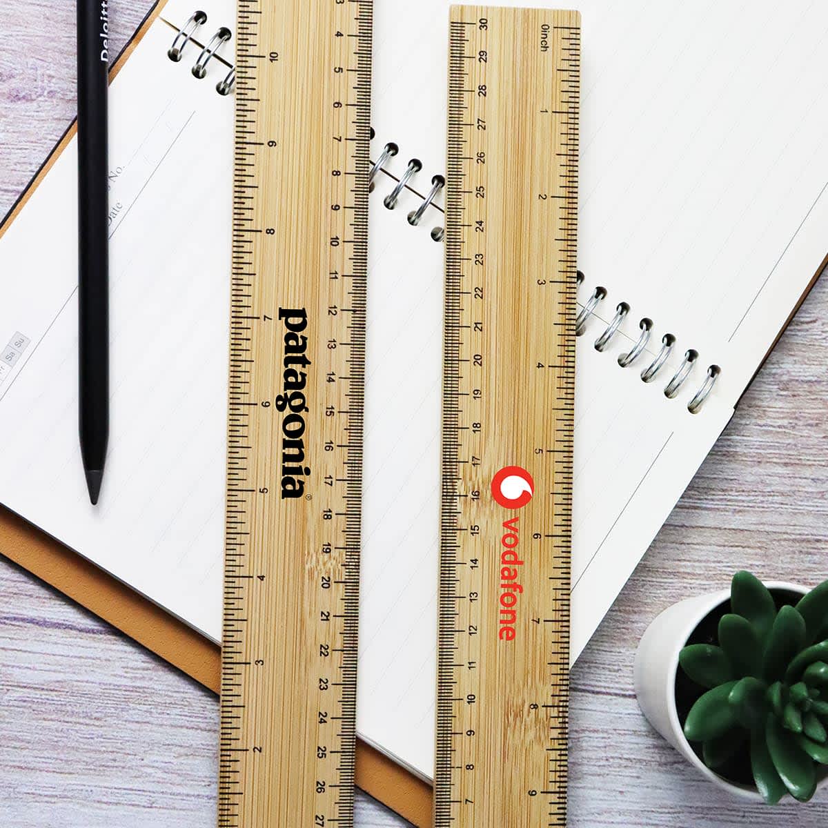 Two bamboo rulers, one with a logo, placed on a notebook with a pen and a small plant nearby.