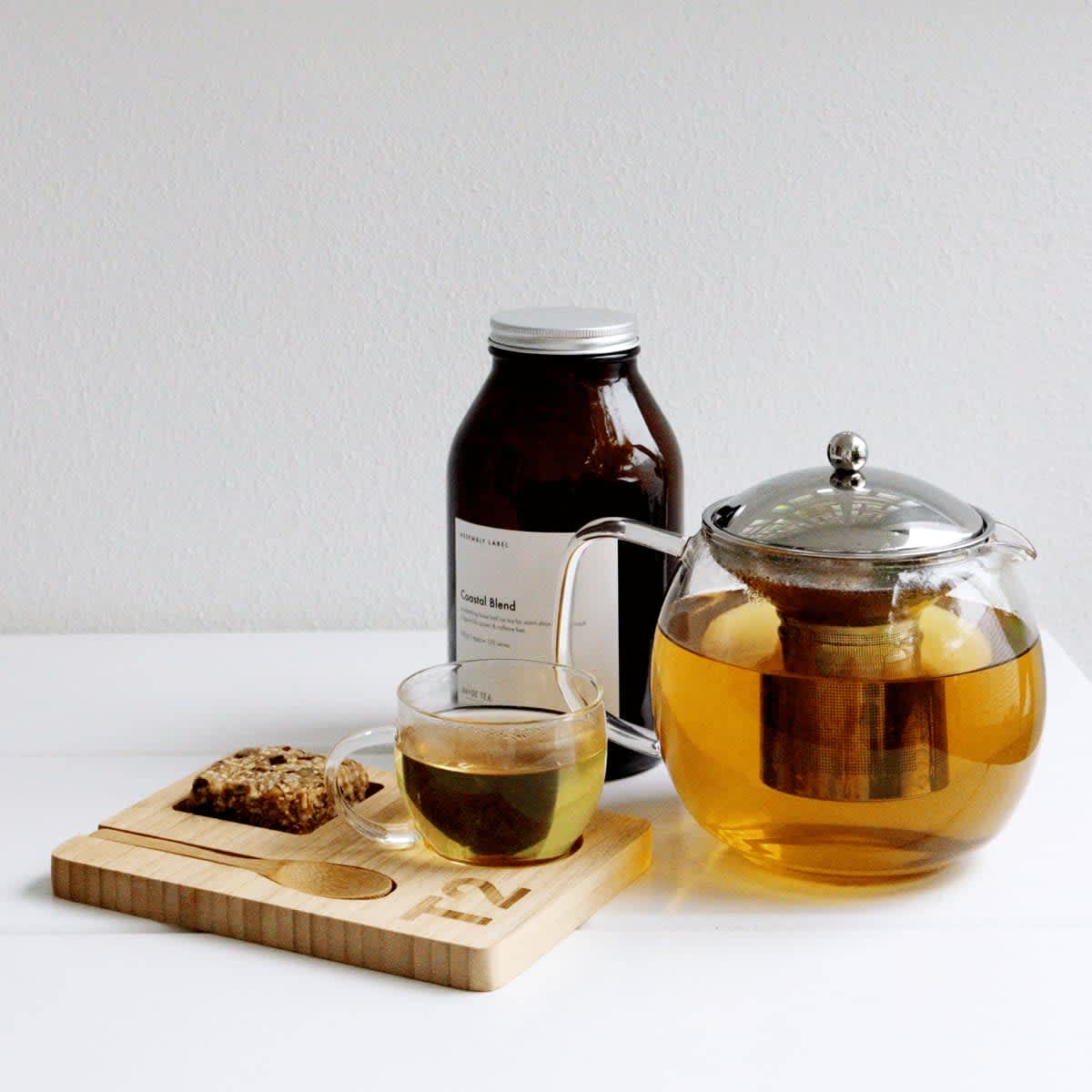 A glass teapot with brewed tea, a cup of tea, a wooden tea tray, and a jar of tea blend. The colours include amber and clear.