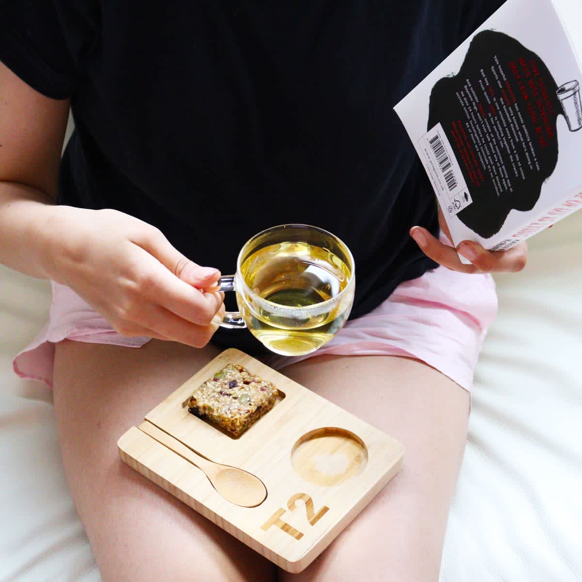 A clear glass mug filled with tea, on a wooden tray with a spoon and a snack, held by a person.