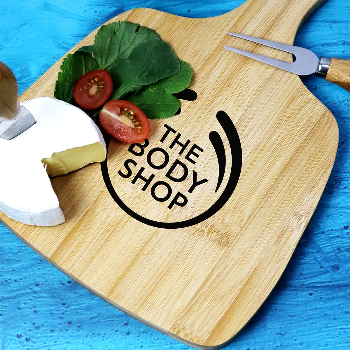 A bamboo cheeseboard featuring a knife, grape tomatoes, and cheese, with a logo on the surface.