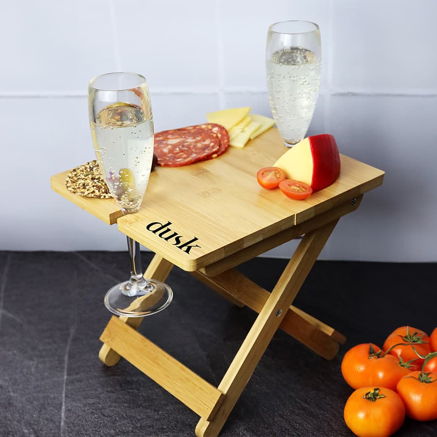 A bamboo folding table with two glasses of sparkling wine, slices of cheese, salami, apple, and cherry tomatoes.