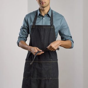 A man wearing a dark denim apron over a light blue shirt, adjusting the apron ties. The apron features a logo.