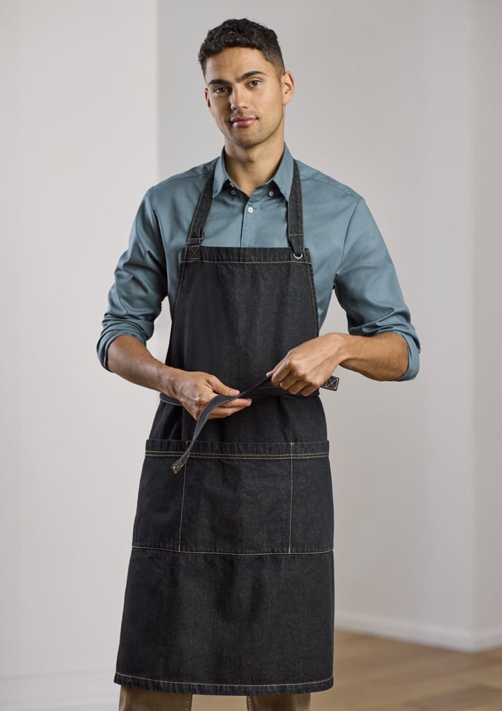 A man wearing a dark denim apron over a light blue shirt, adjusting the apron ties. The apron features a logo.