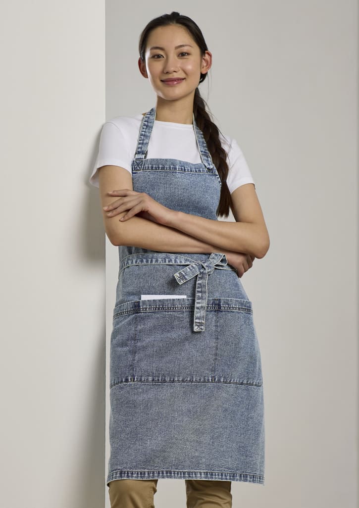 A denim apron in a light blue wash, featuring pockets and an adjustable tie at the waist. The model is smiling.