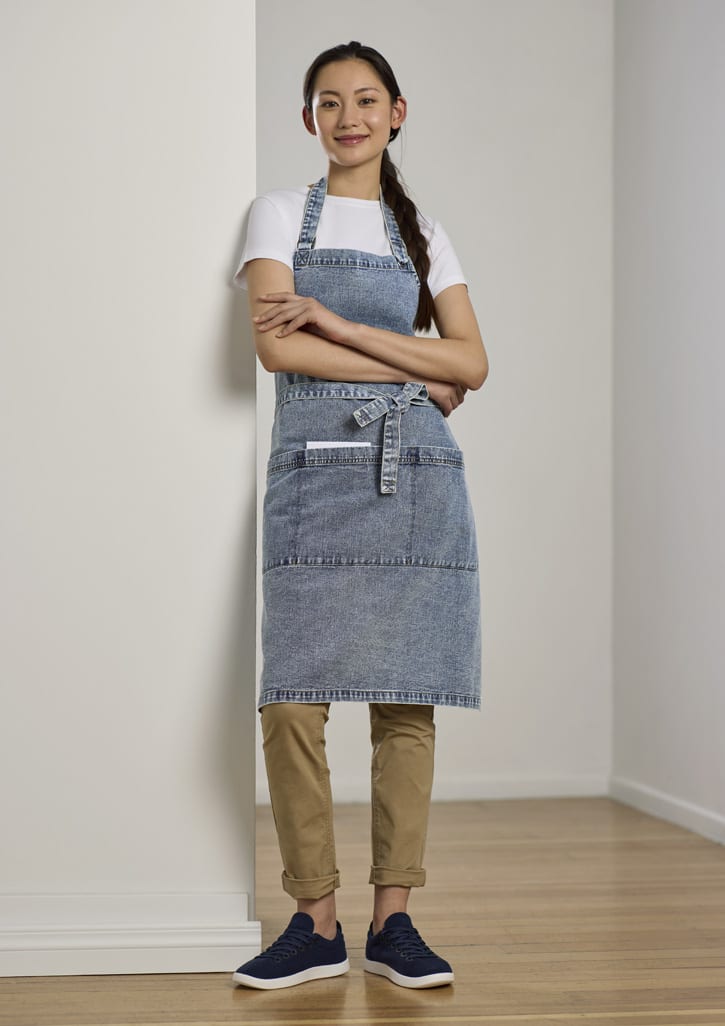 A denim-coloured apron with pockets, worn by a model standing against a wall, paired with a white top and tan trousers.
