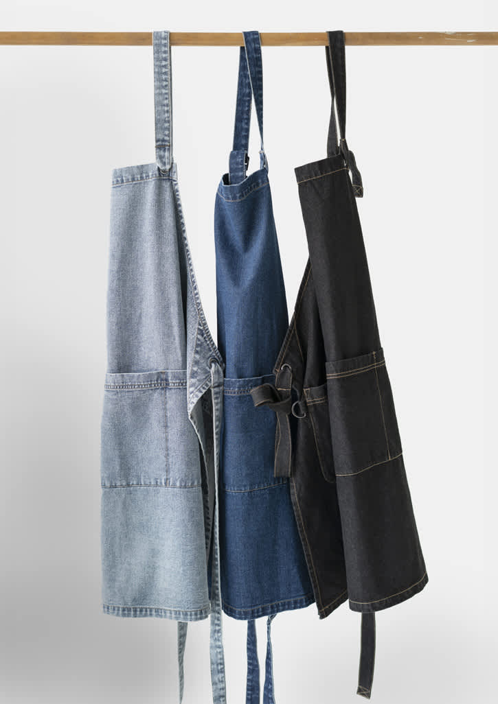 Three denim aprons hanging on a wooden rod, featuring light blue, dark blue, and black colours, each with a pocket and straps.