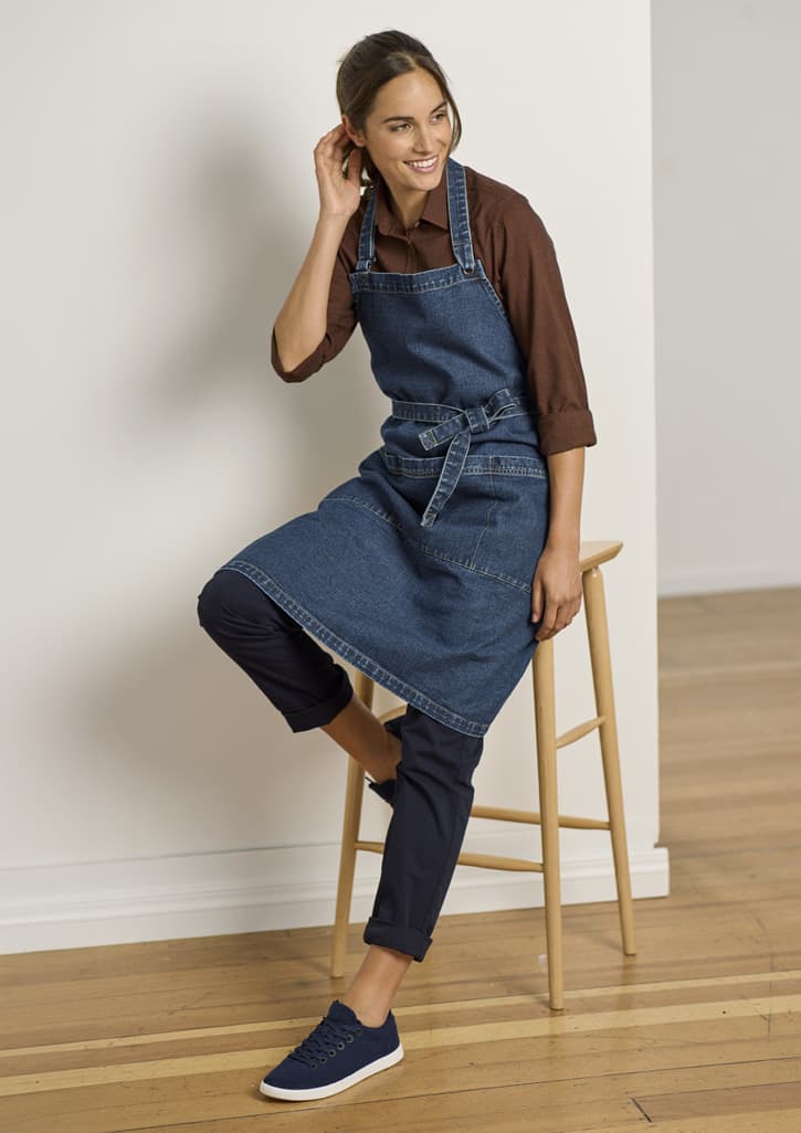 A denim apron worn over a brown shirt, paired with navy pants and sneakers, seated on a wooden stool.
