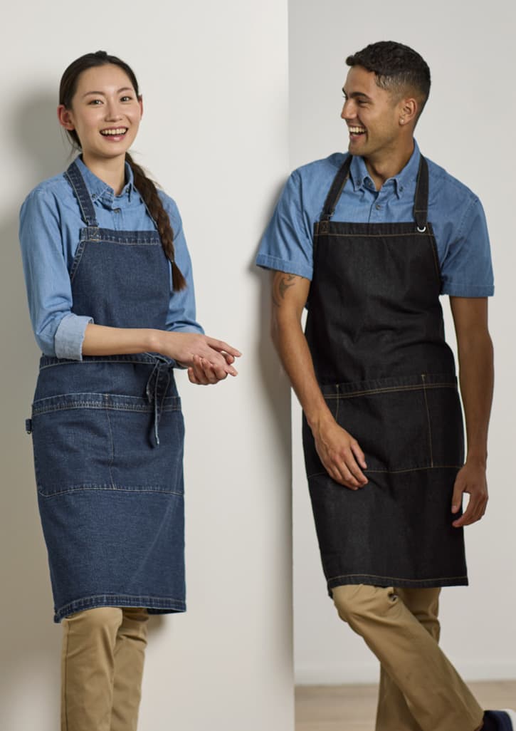 Two people wearing denim aprons, one in a light blue and one in a dark blue, standing against a light wall.