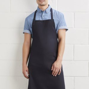 A man wearing a dark navy bib apron over a light blue shirt, standing against a white wall. The apron has a logo.