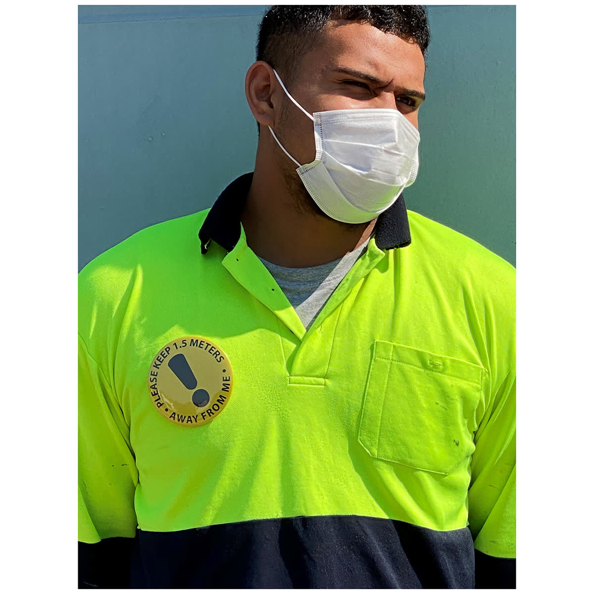 A person wearing a high-visibility yellow and navy polo shirt with a badge and a white face mask.