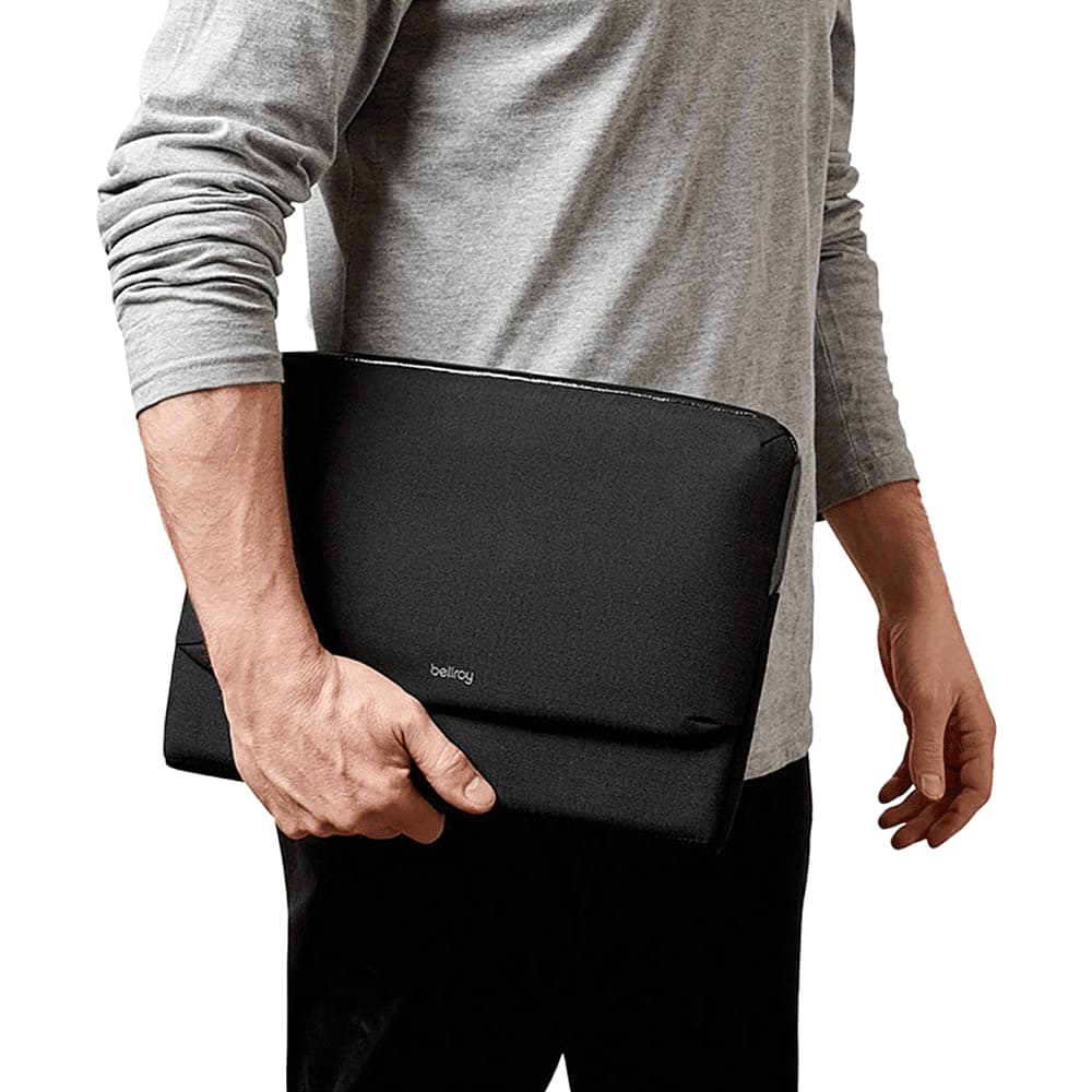 A man in a navy shirt holds a smartphone and a black laptop caddy, standing by a window. The product has a logo.