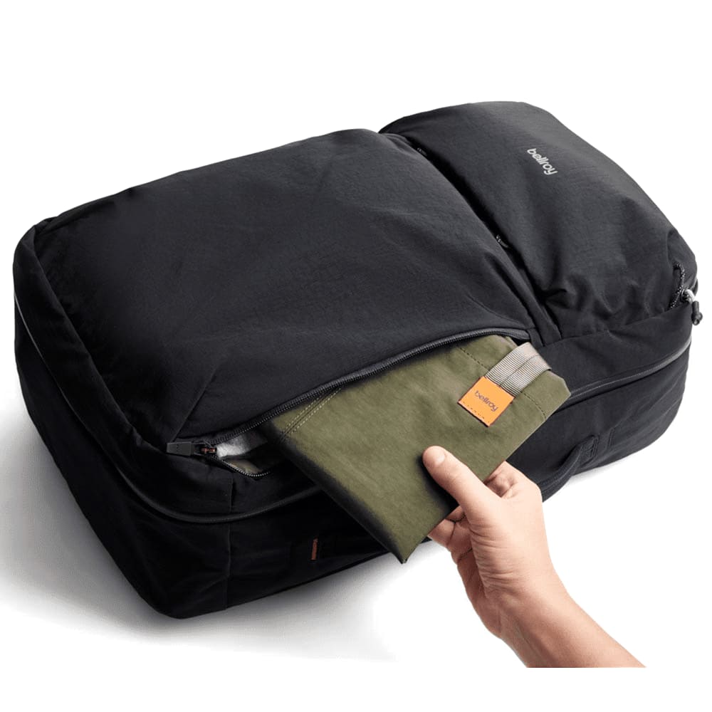 A black travel backpack with a hand reaching into a zippered pocket containing a green pouch.