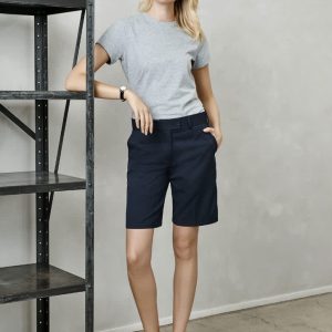 A woman stands beside a shelf wearing navy blue dress shorts and a grey t-shirt, paired with black slip-on shoes.