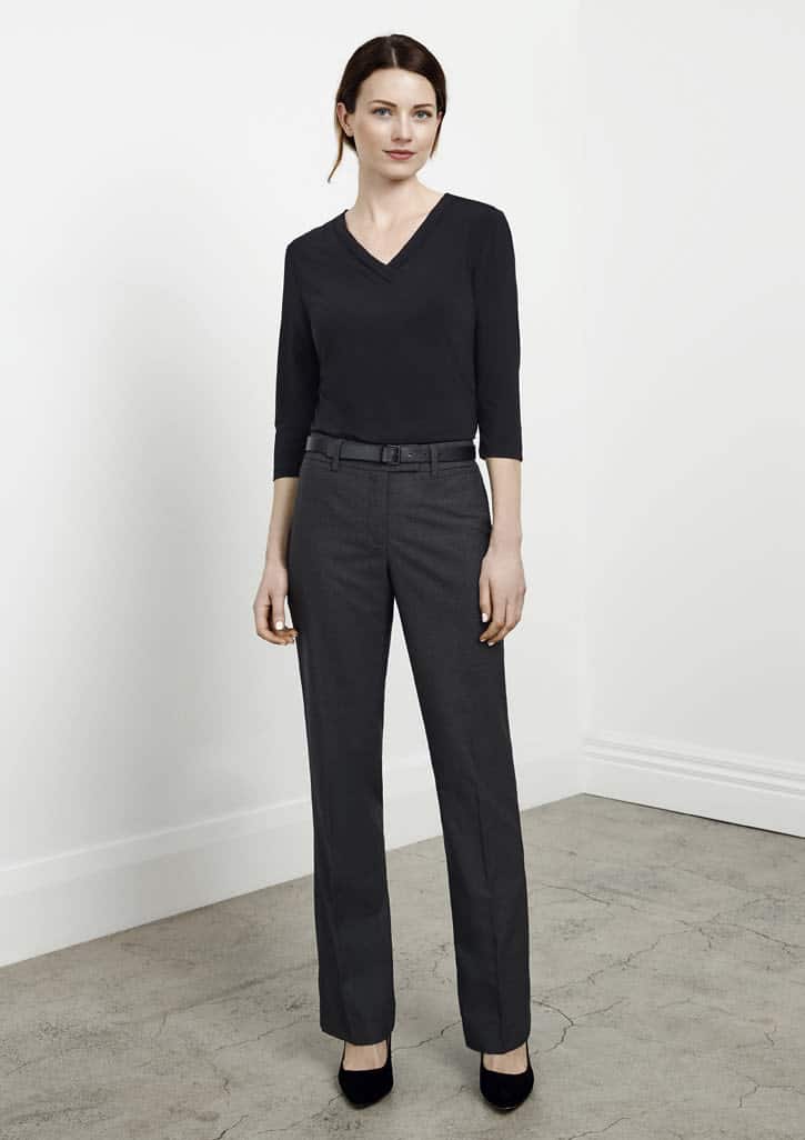 A woman stands wearing black classic pants and a black top, with a belt and stylish heels, set against a light background.