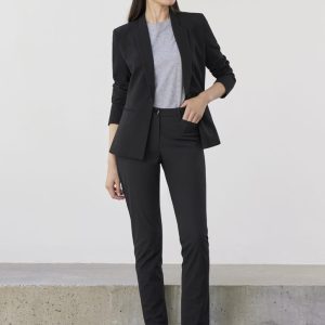 A woman in a black suit with a grey top and black shoes stands against a neutral background. The outfit includes tailored slacks.