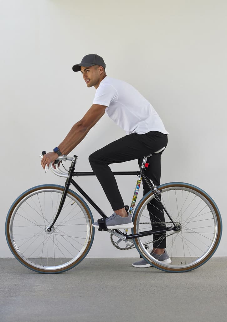 A man in a white t-shirt and black Mens Venture Pants sits on a bicycle, wearing a grey cap and shoes.