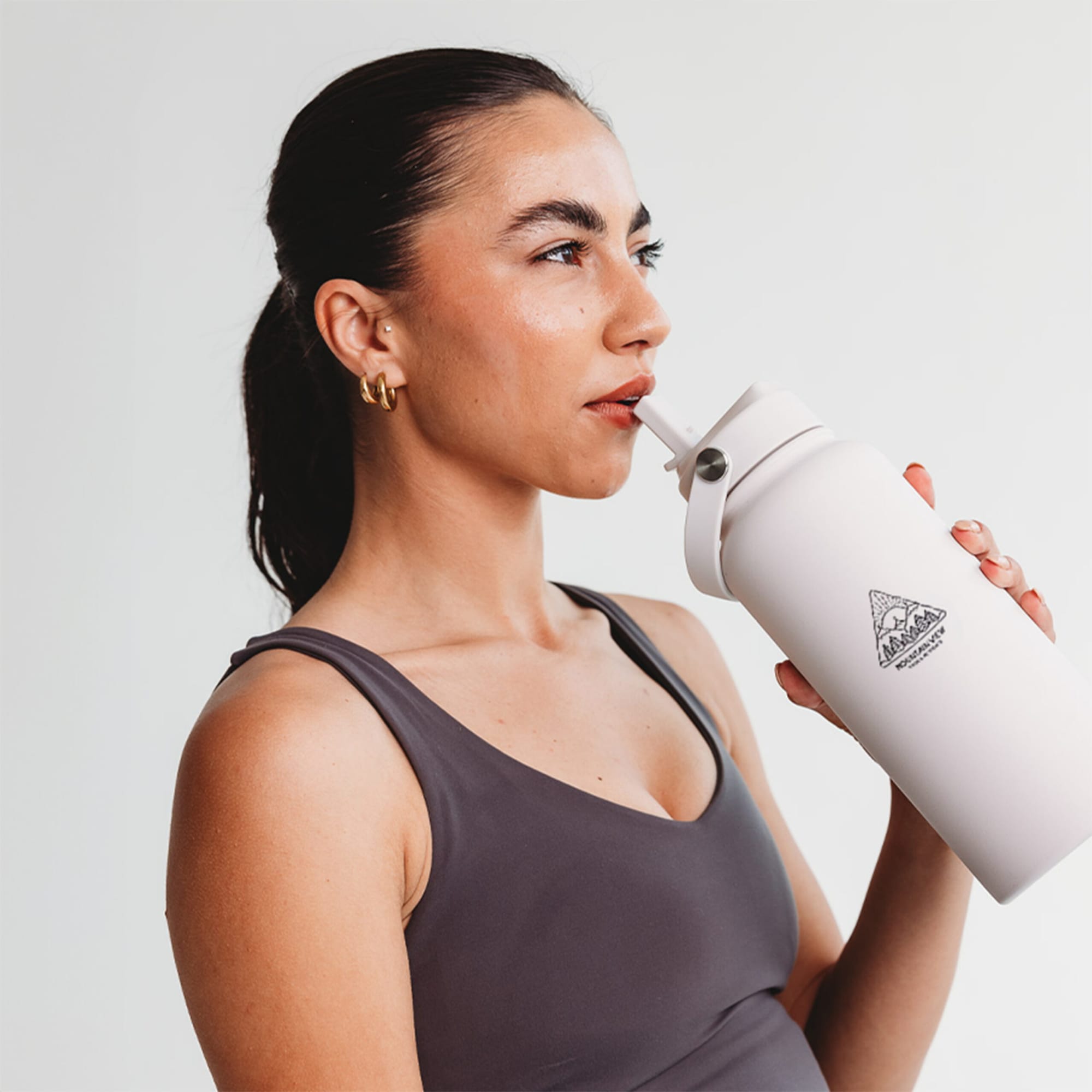 A white drink bottle with a logo, held by a woman in a grey top, sipping through a straw.