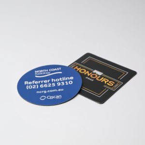 Two round and square coasters, one blue with a logo and contact details, and one black with text, featuring a logo.