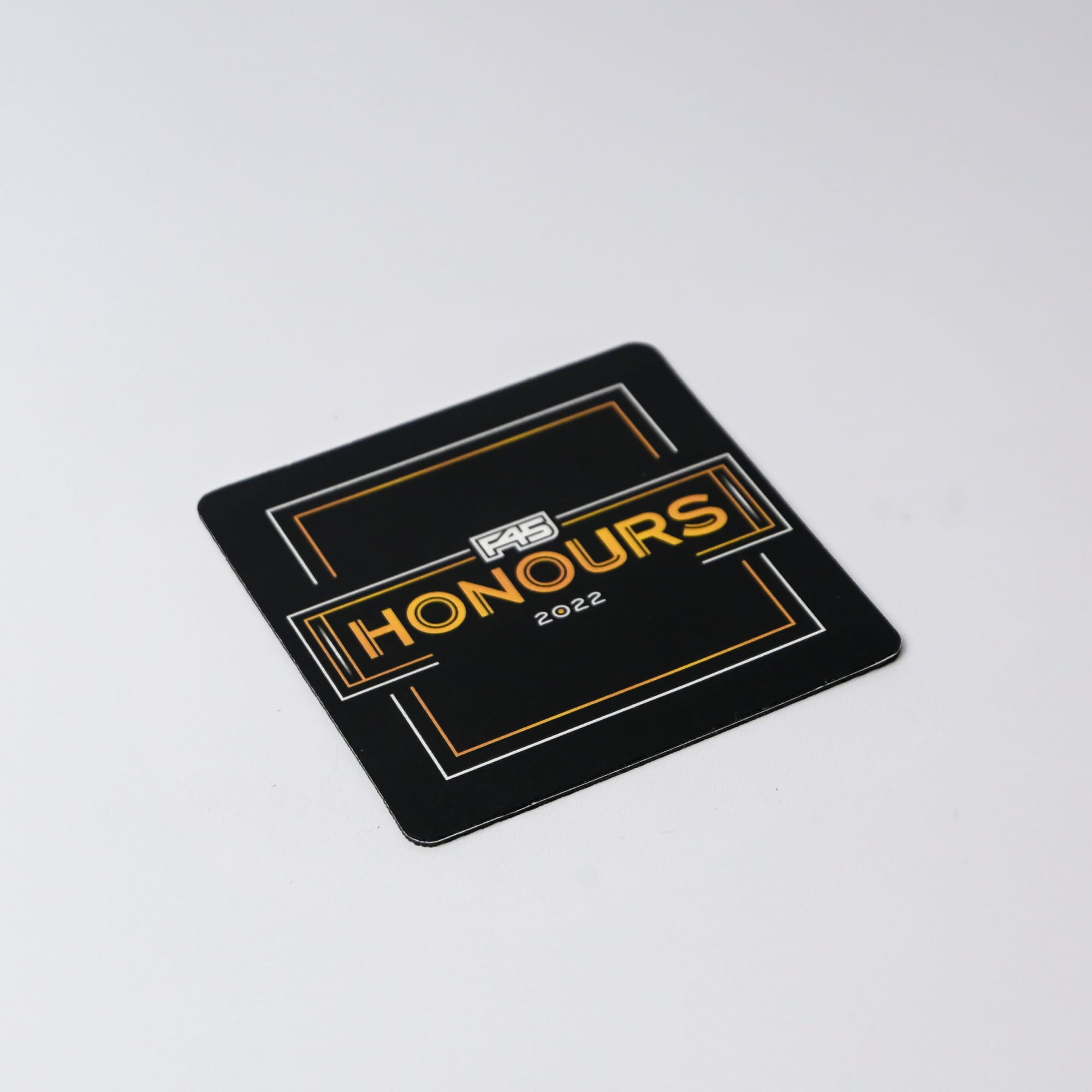 A square black coaster featuring a gold and white logo and the word 