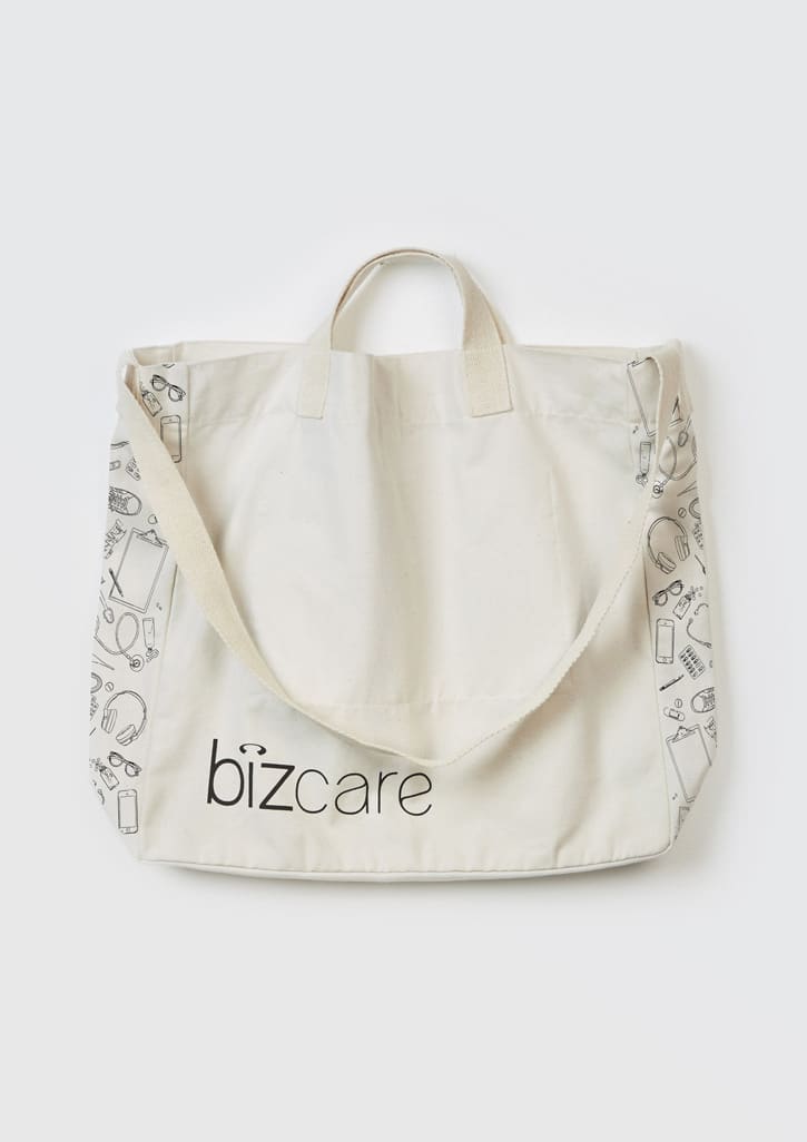 A beige unisex tote bag featuring a repeat pattern of various items and a logo. It has short handles and a long strap.