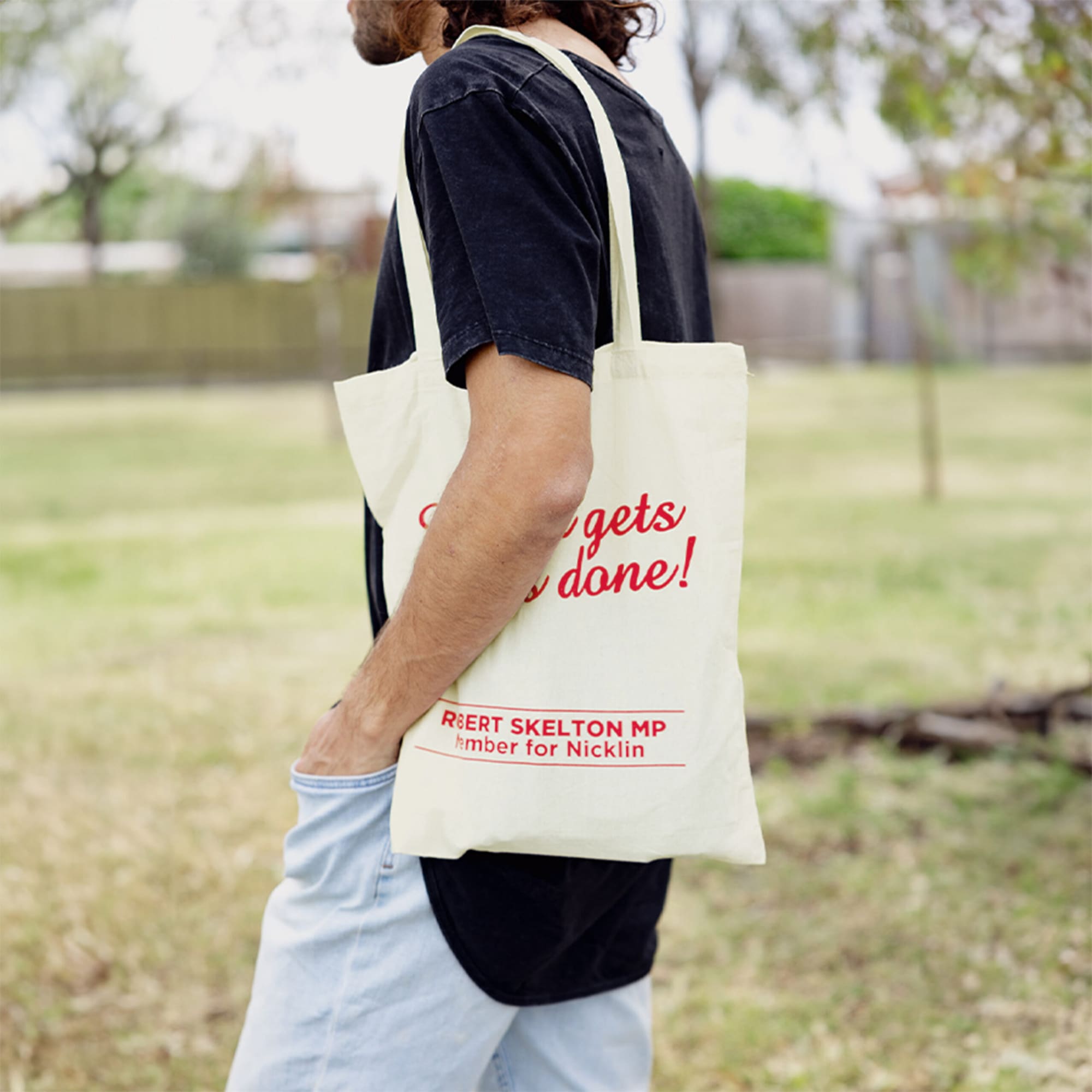 A cream Calico tote bag with a logo, carried by a person in casual attire, against a green outdoor backdrop.