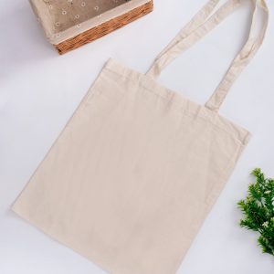 A plain beige tote bag with long handles, placed on a white surface next to a woven basket and some greenery.