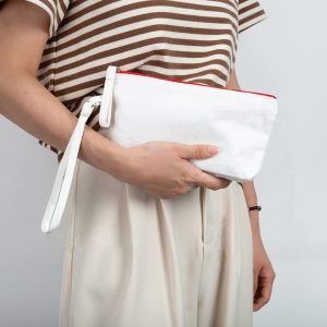A white canvas cosmetic bag with a red zipper, held by a person wearing a striped top and light trousers.